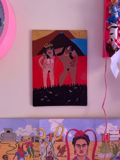 The image displays a collection of art and decorative items arranged on a light-colored wall, likely in an indoor setting. On the left, a large, pink, circular object, possibly a clock face, is partially visible. To its right, a small, rectangular painting depicts two stylized, nude figures against a red background. The figures are outlined in black, with one wearing a gold crown. A dark, textured area representing foliage or ground is at the bottom of the painting. To the right of the painting, a vertical pink border holds several small, toy-like figures, some of which are brightly colored. A thin, pink string hangs vertically in the center of the frame, originating from somewhere above the painting. Below the painting, a panoramic artwork is displayed, featuring various elements of a landscape with architectural structures and stylized figures, including a portrait of Frida Kahlo on the right. The overall scene appears to be a personal display of artistic taste. The image does not contain any identifiable elements that suggest a specific location within Vracar Urban Municipality, Serbia.