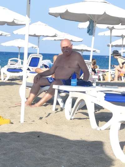 A sunny day at a beach in Lozenets, Bulgaria, features a man sitting on a white beach chair under a white umbrella. He is bald with a mustache and is wearing blue patterned swim trunks. In front of him on the chair is a blue water bottle with the word "lenovo" printed on it. A yellow shoe is visible in the sand to his left. In the background, the blue sea is visible, along with other white beach umbrellas and chairs. Two other people are partially visible in the background.