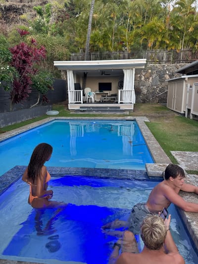 The image displays an outdoor residential backyard scene, consistent with East Honolulu, United States. In the immediate foreground, three individuals are partially submerged in a rectangular hot tub with illuminated blue water. A female with long, dark, wavy hair, wearing an orange bikini, sits with her back to the camera, facing towards the main pool. To her right, a shirtless male with light brown hair and patterned swim trunks leans on the hot tub's edge, holding a dark smartphone in his left hand. In the foreground, a second male, with blonde curly hair and a dark watch on his left wrist, faces away from the camera, holding a smartphone.
The hot tub is integrated with a larger, clear blue swimming pool. Beyond the pool, an open-sided cabana or pool house, painted white with white railings and curtains, stands on a grassy area. Inside the cabana, a television is mounted on the back wall, along with a white plastic chair, a table, and what appears to be exercise equipment. To the right of the cabana, a rock retaining wall and three light-colored storage sheds are visible. To the left of the pool, a black retaining wall supports red-leafed plants and other green shrubs. A deflated inflatable ring lies on the grass near the black wall. The background features a dense, lush hillside covered with tropical vegetation, including numerous palm trees, situated above a wooden fence.