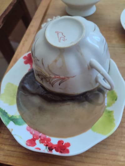 A white teacup, decorated with delicate brown and red floral patterns, is overturned on its saucer. The saucer is also white and adorned with similar floral designs, featuring red berries and green leaves. A dark liquid, likely coffee or tea, has spilled from the cup and pooled on the saucer, creating a glossy, reflective surface. The cup itself has a small red emblem on its underside. The scene is set on a wooden surface, possibly a table, with hints of other objects in the background, including what appears to be another white porcelain dish and a portion of a wooden structure. The lighting suggests it is daytime, and the overall impression is one of a domestic or cafe setting. No specific time of day, weather, or emotional cues are discernible from the image. The only visible text is the red emblem on the teacup.