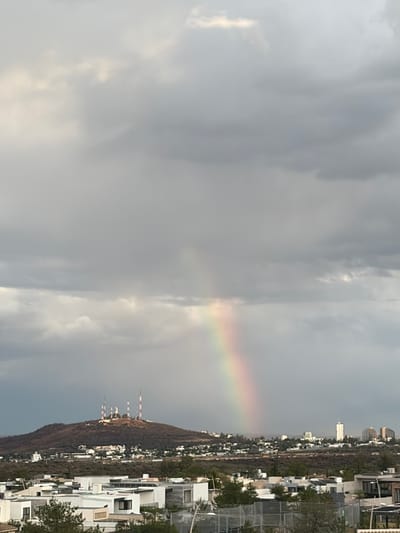 The image captures a panoramic view of an urban landscape set against a hilly backdrop under a cloudy sky. The primary subject is a vibrant, albeit partial, rainbow arching across the central sky, contrasting with the predominantly grey and white overcast clouds.
On the right side of the frame, modern, mostly white residential buildings with contemporary architecture are visible, extending towards the base of a large hill or mountain. These structures appear to be part of a developed residential area, with some green trees and shrubs interspersed.
The left side of the image is dominated by a sprawling hill or mountain, covered in what appears to be dry or sparse vegetation. Along the crest of this hill, several tall, slender structures, likely communication towers or antennas, stand out, some featuring distinctive red and white markings. In the distant background, beyond the hill, further urban development is visible, including taller buildings and a faint city skyline.
The weather is characterized by heavy cloud cover, with pockets of brighter light suggesting the sun is breaking through, which is conducive to the formation of the visible rainbow. The time of day appears to be during a period of mixed weather, possibly after a rain shower in the late afternoon. There are no people visible, and no discernible text. The scene is consistent with a city like León, Mexico, known for its mountainous surroundings and urban development.
