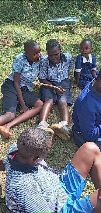 The image shows five young boys seated in a grassy area. They are in a playful mood, with one boy holding a stick in his hands. Three of the boys are wearing blue and white checkered shirts and blue shorts. One boy is wearing a blue shirt and navy blue shorts. Another boy is wearing a blue shirt and navy blue shorts. The boys are interacting and laughing. The location is Bungoma, Kenya, with coordinates 0.553 latitude and 34.773 longitude. The background is a lush green lawn. The sun is shining and the sky is blue.