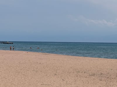 The image depicts a serene beach scene on a cloudy day, likely in the afternoon, at Kara-Oy, Kyrgyzstan. The foreground is dominated by a wide expanse of light brown sand, showing sparse patches of dry grass. The sand meets the calm, deep blue water of a lake or sea.

In the middle ground, several people are visible. On the shore, two individuals stand near the water's edge, one wearing a light-colored top and shorts, the other in dark swimwear. Further out in the water, a group of people are swimming and enjoying the cool blue water, with their heads and shoulders visible above the surface. The water itself has gentle ripples, suggesting a calm atmosphere.

In the far background, a long, wooden pier extends into the water, with a few more indistinct figures standing on it. Beyond the water, a faint, hazy mountain range is visible on the horizon, partially obscured by the cloudy sky. The sky is a muted, pale blue with streaks of white clouds, indicating overcast weather. The overall mood is peaceful and leisurely, capturing a typical summer day at a lakeside or seaside resort. There is no visible text in the image.