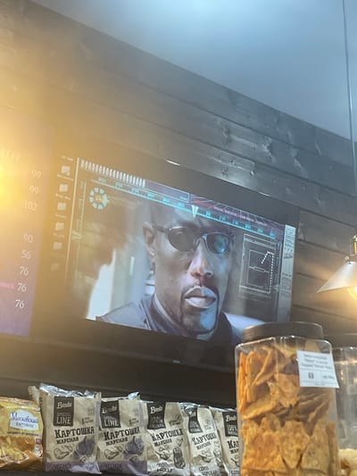 This media file shows a close-up of a television screen displaying a scene from a movie or video game. The screen shows a person's face, partially obscured by glasses, with a futuristic heads-up display overlaying the image. The display includes various graphical elements, text, and icons, suggesting a sci-fi or action genre. To the right of the television screen, shelves are visible, stocked with packaged potato chips. The packaging features text in Russian, including brand names like "Bruto" and descriptions such as "КАРТОШКА ЖАРЕНАЯ" (fried potatoes) and flavors like "ЧЕСНОК И ТРАВЫ" (garlic and herbs). The overall setting appears to be an indoor environment, possibly a store or a home entertainment area, given the presence of merchandise. The lighting is somewhat dim, with a bright light source visible at the top of the frame, potentially a lamp or overhead light. The image is slightly tilted, suggesting it was taken at an angle.