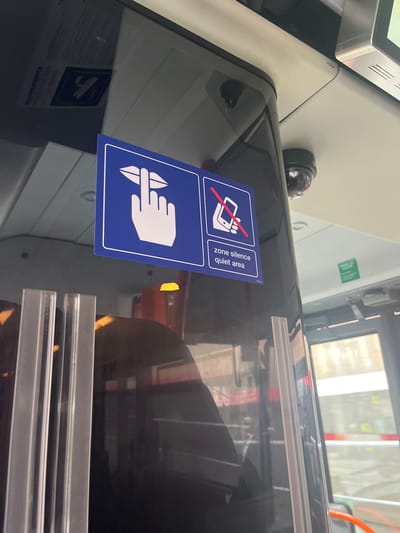 This image captures a detail inside what appears to be a public transport vehicle, likely a tram or bus, in Genève, Switzerland. The primary subject is a blue and white rectangular sign affixed to a reflective, dark surface, possibly a window or partition within the vehicle.
The sign features two distinct pictograms and accompanying text, indicating a designated "quiet area." The upper section shows a white mobile phone with a red circle and a diagonal line through it, universally understood as a prohibition against using mobile phones, specifically for calls. Below it, a white hand with an index finger pressed to the lips signifies a request for silence. The text "zone silence quiet area" is written vertically in white letters alongside these symbols.
Other objects visible in the scene include a small, black dome-shaped security camera mounted above the reflective surface. Reflections on this surface distort parts of the interior and what seems to be the exterior through a window, showing vague shapes and light. In the lower left of the reflective surface, another, less clear sign with a faint airplane icon can be glimpsed in reflection. Metallic strips or frames are visible on the right side, further suggesting the interior structure of a vehicle.
The overall impression is of an area designated for passenger tranquility, with rules enforced through signage. The lighting suggests it is daytime, with light filtering in from outside the vehicle. No people are directly visible in the frame, though their presence is implied by the setting of public transport.