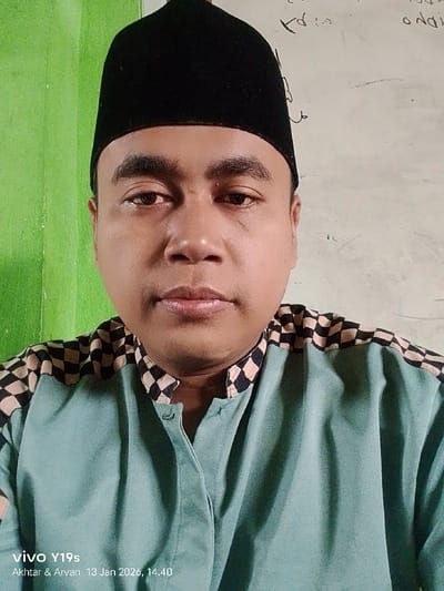 This is a close-up, head-and-shoulders portrait of a man, likely of Southeast Asian descent, appearing to be in his 30s or 40s. He is looking directly at the camera with a neutral, composed expression. He wears a black songkok (a traditional cap) and a light teal or blue long-sleeved shirt with a distinctive black and light beige checkered pattern on the shoulders and collar. The shirt has a band collar and a partial button placket.
The setting is indoors, with a bright green wall visible to the viewer's left (behind the man's right shoulder). To the viewer's right, behind his left shoulder, is a white surface that appears to be a whiteboard with faint, illegible handwritten script on it, suggesting a classroom or office environment.
The man is posing for a photograph, likely a selfie. A digital watermark in the bottom left corner provides details: "vivo Y19s" (indicating the smartphone model), and "Akhtar & Arvan 13 Jan 2026, 14.40". This timestamp indicates the photo was taken at 2:40 PM on January 13, 2026. The location of the scene is Wanguk, Indonesia. The lighting is even, suggesting an indoor light source.