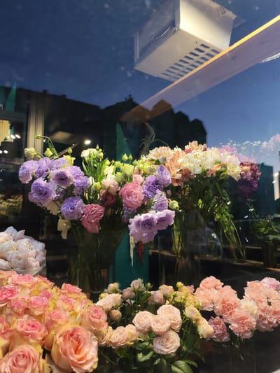 The image depicts a flower shop window display in Nizhnevartovsk, Russia. In the foreground, a variety of flowers are arranged in vases. These include bouquets of purple and pink lisianthus, a bunch of light pink roses with yellow centers, and a cluster of pale pink carnations. The flowers are presented in clear glass vases, with some of their stems visible. Behind the flowers, a reflection of the sky and buildings is visible in the window. The sky is a deep blue, suggesting it is either dusk or dawn. The buildings in the reflection appear to be dimly lit. A white rectangular fixture with a grated surface is also reflected from above. The overall scene is illuminated by the soft light of the sky and possibly interior lighting from the shop, creating a serene and colorful display.