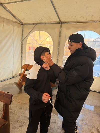 Two young males are inside a tent-like structure, possibly in Zlatna Panega, Bulgaria. One male, wearing a black hooded sweatshirt with a white panel, holds a lit cigarette in his right hand and has his left hand raised to his face. He appears to be sitting on a wooden bench. The second male, wearing a black winter coat and a black baseball cap, stands over the first male, and his right hand is on or near the first male's face. The interior of the tent is visible, revealing a metal frame and translucent fabric. Outside the tent, there is the vague image of a building. The floor inside the tent appears to be concrete.