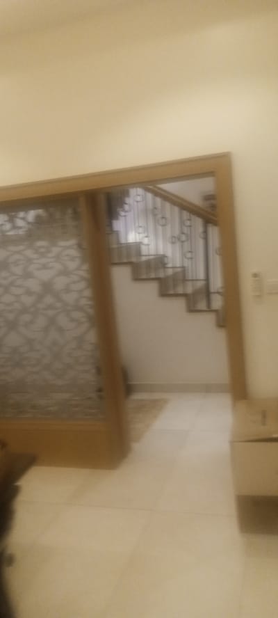 An interior shot appears to show an entranceway with light wood trim, possibly leading to a staircase. The staircase has decorative metal railings and is visible in the background. A patterned screen or partial wall is on the left. The flooring appears to be light-colored tile. A piece of furniture is visible on the right. No people are visible. The specific location in Unguwar Rimi, Nigeria cannot be determined from the image.