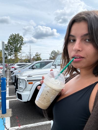 A young adult female is depicted in the foreground, holding a Starbucks beverage in an outdoor parking lot in West Windsor, United States. She has dark brown hair and is wearing a dark tank top beneath a dark brown cardigan; a small tattoo is visible on her upper left shoulder. She holds a clear plastic Starbucks cup containing a light-brown blended drink topped with whipped cream, with a green plastic straw positioned between her lips, and is looking directly at the viewer.
The background features a paved parking lot with multiple vehicles. A white Mercedes-Benz G-Wagen is prominently parked directly behind the female, followed by a row of other light-colored SUVs and sedans. A blue parking bollard stands to the left. The environment is under a partly cloudy sky, with green trees and utility poles visible in the distance, indicating a commercial or retail area.