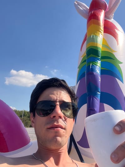 A selfie, taken in the outdoors, shows a man wearing sunglasses, holding a white styrofoam cup and positioned with a large inflatable rainbow unicorn above him and a pink inflatable tube below. The background shows a clear blue sky with sparse clouds and some trees in the distance. The photograph seems to be taken during the day, under bright sunlight, potentially in San Fernando, Argentina.