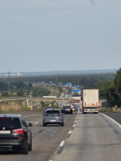 The image depicts a highway scene in Germany, with a focus on a traffic jam. Multiple vehicles, including cars and trucks, are visible on the asphalt road. The sky is overcast. In the distance, a dense forest borders the highway, with some industrial structures and power lines visible on the horizon. A concrete overpass crosses the highway further up ahead. The scene is set in Wicker, Germany.