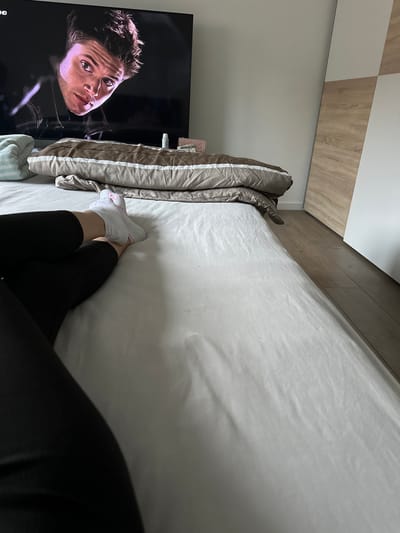 A person is lying on a bed, with their legs crossed and wearing white socks and black leggings. The bed is made with a white sheet and two pillows are visible. In the background, a large television screen shows a close-up of a man's face. A wardrobe with wooden and white doors is partially visible to the right of the bed. The scene appears to be in a bedroom.