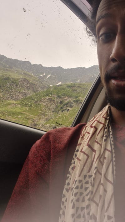 A selfie taken from inside a vehicle shows a person's right side of their face and upper body against a backdrop of a mountain landscape seen through a rain-streaked window. The person has dark curly hair and a mustache, and is wearing a red garment and a patterned scarf. The mountains are green and rocky, with patches of snow visible. A "SUZUKI TEMPERLITE" sticker is visible on the window. The scene suggests travel in a mountainous region of India during overcast weather.