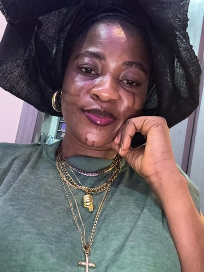 A portrait-style, close-up photograph shows a Black woman wearing a black headscarf and a green t-shirt. She is resting her chin on her hand, with her fingers gently touching her cheek. Her eyes are open and she appears to be looking towards the camera. She is wearing multiple layered gold necklaces, one of which has a crucifix pendant with clear gemstones. She also has a gold hoop earring in her visible ear. Her lips are colored with a glossy, bright pink lipstick. The lighting in the image is somewhat diffused, creating a subtle sheen on her skin. The background appears to be indoors, with a blurred hint of shelving or furniture. The overall mood of the image is intimate and perhaps contemplative.