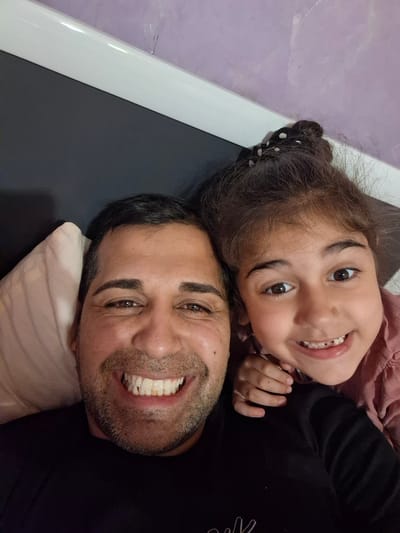 An overhead, slightly rotated shot captures a man and a young girl lying down together, smiling broadly at the camera. The man, positioned higher in the frame, has dark hair, stubble, and is wearing a dark top. A light-colored pillow is visible behind his head. He has a wide, joyful smile, revealing his teeth.

Below him, to the right, is a young girl with dark, curly hair tied back with a black scrunchie featuring white dots. She is also smiling widely, showing her teeth, and her eyes are looking directly at the viewer. Her left hand is resting near the man's chest. She appears to be wearing a dark long-sleeved top over a pink or mauve garment.

The background suggests an indoor setting. To the left of the man, a dark, flat surface is visible, possibly part of a bed frame or furniture. To the right and below the girl, a light purple wall is visible, separated by a thin, white vertical element, likely part of the furniture or room decor. Both subjects appear relaxed and happy, conveying a warm and affectionate moment. There is no visible text in the image. The scene takes place in Novachene, Bulgaria.