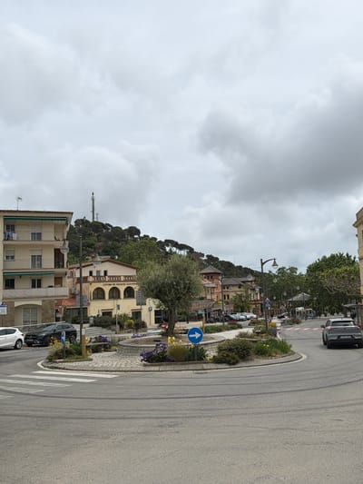 The scene shows a street in Sant Andreu de Llavaneres, Spain, featuring a roundabout with a blue directional arrow sign. A white pedestrian crossing is visible near the roundabout. Buildings are situated on a hill in the background, partially obscured by trees. Several cars are parked alongside the road and some are moving. A telecommunications tower is visible in the distance. The sky is overcast with grey clouds.