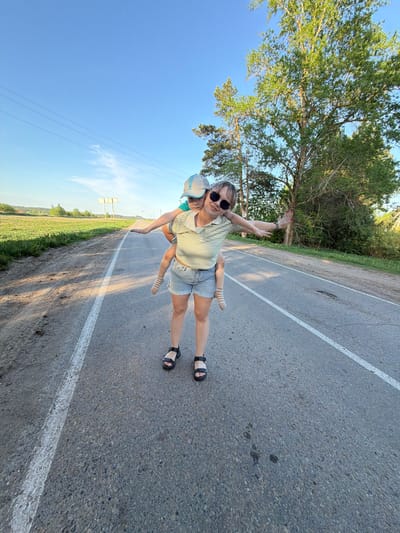 A light-skinned woman is giving a light-skinned child a piggyback ride on a paved road, flanked by green grass on one side and green foliage with trees on the other side. The woman is wearing dark sunglasses, a light-colored striped short-sleeved shirt, and jean shorts with black sandals. The child is wearing a hat and a light-colored shirt and striped socks with black sandals. A white painted line runs along the side of the road. The sky is blue. The location cannot be confirmed as Foki, Russia from the provided image.