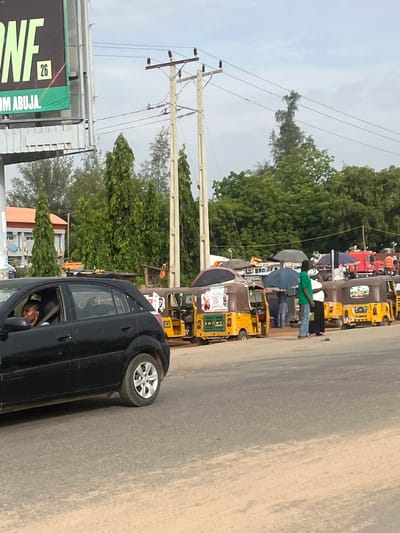 The image shows a street scene in Galadimawa, Nigeria. On the left side, there is a black car and a sign that reads "26" and "UM ABUJA". A person is visible inside the car. Along the roadside, there are numerous yellow auto-rickshaws parked. Several people are standing or walking near the auto-rickshaws. The background includes trees and power lines against a cloudy sky. The road surface is light-colored.