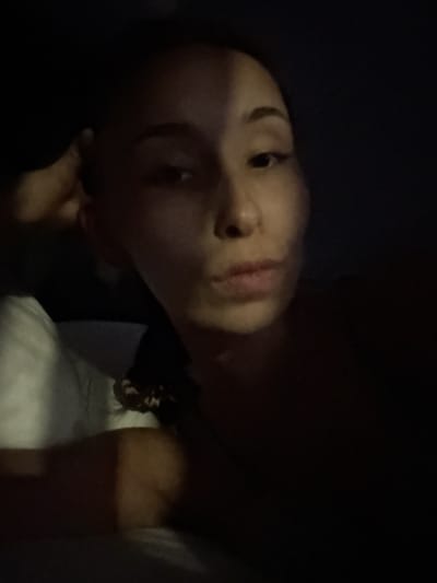 The image shows a close-up, low-light selfie of a person's face. The lighting creates a stark shadow diagonally across the face, obscuring the left side of the forehead and eye. The right side of the face is more illuminated, revealing the right eye, the bridge of the nose, and the mouth. The person appears to be lying down, with a white pillow visible on the left side of the frame. The background is dark and indistinct. The location, Doma 1096 Km, Russia, is a geographical marker, but the visual information in the image does not provide any specific clues about this location.