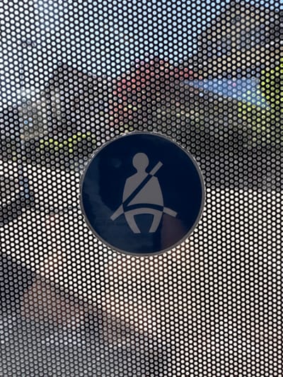 The image displays a circular blue and white sign depicting a person wearing a seatbelt, indicating a safety regulation. This sign is affixed to a perforated surface, possibly a window or screen, with a bokeh effect in the background showing out-of-focus greenery and hints of red and blue, suggestive of an outdoor urban environment. The image is taken at a slight angle, with the sign centrally located and partially obscured by the perforations. The scene does not provide specific visual cues that would allow for precise location identification within Reconvilier, Switzerland.