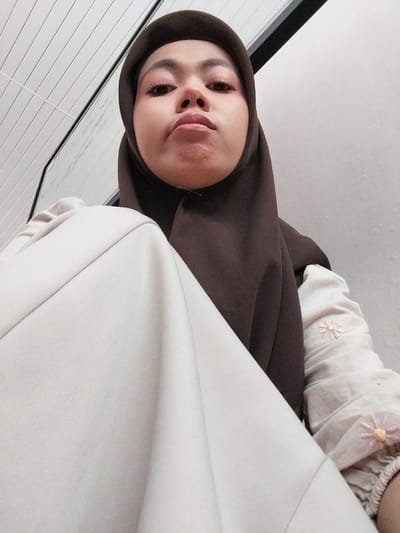A selfie is captured from a low angle, featuring a person wearing a dark brown hijab and a light-colored shirt with embroidered pink and yellow flowers. The person's face is tilted upwards, with their eyes looking directly at the camera. Their lips are slightly parted, and they appear to have makeup on, including eyeliner and mascara. The background includes a white, slatted surface on the left, which could be part of a window blind or wall paneling, and a white fabric that drapes across the right side of the frame. The lighting suggests it might be daytime, possibly indoors given the artificial look of the slatted surface. The overall mood is casual and personal, typical of a selfie. There is no visible text in the image. The location is identified as Tibojong, Indonesia.