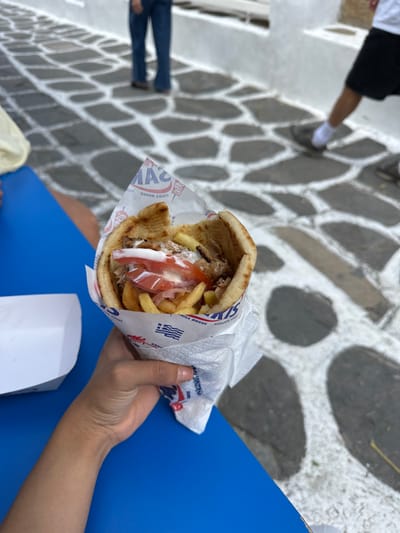 A close-up, slightly elevated perspective image depicts a hand holding a Greek gyros in a pita bread, centrally positioned in the lower half of the frame. The gyros is wrapped in white paper featuring blue and red text including "SAM'S GRILL HOUSE" and "MYKONOS GREECE." The pita is filled with vertical slices of roasted meat (likely pork or chicken), french fries, red tomato slices, and a white creamy sauce. The hand is positioned over a bright blue table or surface. To the left of the gyros, a white, empty paper tray rests on the blue surface. The background reveals a distinct street pavement composed of dark grey, irregular cobblestones separated by wide, white grout lines, a pattern typical of Mykonos, Greece. White walls are visible flanking the street. Blurred partial figures of at least two individuals are in the background, walking away from the camera: one wearing blue jeans and brown shoes, and another wearing black shorts, white socks, and grey sneakers. A third person's upper arm/shoulder, clothed in light yellow fabric, is partially visible on the far left edge, near the blue table. The scene occurs outdoors during daylight.