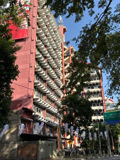 The image shows a medium-shot view of a building in Colombo, Sri Lanka. The building has a distinctive red exterior with rows of projecting white structures, which are likely balconies or sunshades. Trees partially obscure the view of the building, and other taller buildings are visible in the background. A street sign is visible, along with decorations and streetlights lining the street. The sky is partially visible at the top of the image. Overall, the image presents a typical urban scene in Colombo.