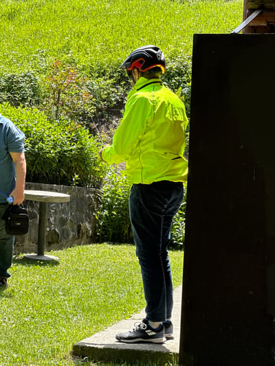 The image depicts two individuals outdoors, likely in a park or garden setting, with a backdrop of lush green grass and foliage. The primary subject, positioned on the right, is a person wearing a bright yellow, long-sleeved jacket and dark blue jeans, along with a black and red cycling helmet. This individual is standing next to a dark, vertical structure, partially obscuring their right side. Their stance suggests they are facing away from the camera, looking towards the left.

To the left of the frame, another person is visible from the waist down, wearing a blue shirt and dark jeans. They are holding a black messenger bag in their left hand and appear to be standing near a round, concrete table supported by a stone base. This individual is facing towards the right.

The environment suggests a sunny day, with dappled light illuminating the grassy areas and plants. The presence of the cycling helmet on the main subject could indicate an activity related to cycling. The concrete table and stone wall suggest a possibly maintained outdoor area. The overall scene conveys a casual, outdoor gathering or moment. The specific location within Bare Kraljske, Montenegro, is not explicitly discernible from the visual elements present.