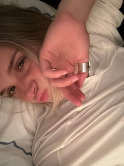 A woman is lying on a bed in an indoor setting. She is fair-skinned, with blonde hair and wearing a silver ring on her right hand. She is looking at the camera. Her hand is partially obscuring her face. She has a small ring in her nostril. She is lying on a bed with white sheets and blankets. The location in the city of Whistler, Canada cannot be determined from the image.