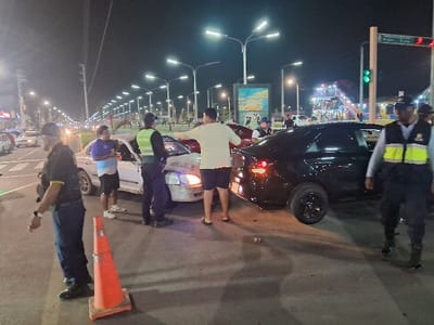 The image depicts a nighttime road scene in the city of Nuevo Chimbote, Peru, at what appears to be the aftermath of a traffic incident.
Two sedans are centrally positioned on a multi-lane asphalt road. A white four-door sedan exhibits significant frontal damage, with its hood crumpled and bumper displaced. Its headlights are illuminated. This vehicle is positioned diagonally across a lane. Behind it, a black four-door sedan is situated slightly to the right, its rear lights illuminated.
Six individuals are visible in the scene. Three are uniformed personnel: one on the far left, facing right, wearing a dark cap and dark uniform with a yellow arm stripe; one in the center-left, facing the white car, wearing a dark uniform with a bright yellow-green reflective vest; and one on the far right, wearing a similar uniform and vest, walking towards the foreground. Three civilians are present: one male near the driver's side of the white car, wearing a purple and white shirt, dark shorts, and white sneakers, holding a can or bottle; one male in a light-colored shirt and dark shorts, positioned between the two vehicles, gesturing towards the white car; and another individual partially visible in the driver's seat of the black car.
An orange traffic cone with a white base rests on the asphalt in the immediate foreground. Road markings, including white lines and a green-painted lane demarcation, are visible. Small pieces of debris are scattered on the road near the front of the white car.
The environment is illuminated by numerous tall streetlights with bright, modern fixtures lining the road. Traffic lights are visible in the distance, with one showing a green signal. Commercial buildings or storefronts with some visible signage are present along the left side of the road. In the far background to the right, a brightly lit amusement park or fairground, featuring visible rides and structures, is prominent. A large billboard or digital display screen is also visible above the black car.