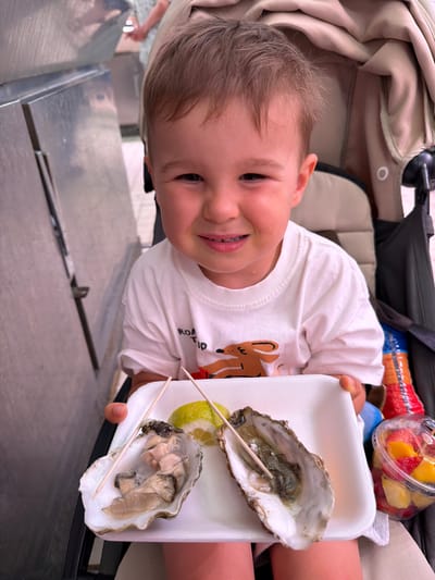 A young child, likely a toddler, smiles with teeth showing while lying on their back in a stroller. The child is wearing a white t-shirt with a deer graphic. The child is looking up towards the camera and appears to be happy. The child is holding a white Styrofoam tray with two oysters and a slice of lemon. The oysters are open and appear to have been recently served. In the background, a clear plastic container filled with fruit salad is visible, suggesting a casual outdoor meal. The overall setting appears to be a public area, possibly a park or outdoor seating area, given the stroller and the presence of other people in the background. The lighting suggests it could be daytime. The child's expression conveys a sense of joy and curiosity. The text "ROAD TRIP" is visible on the child's t-shirt. The scene is likely taking place in Alboraia, Spain.