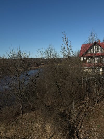The photograph depicts a building in the vicinity of Колчаново, Russia, as discerned from the architectural style and surrounding environment. The building has a red roof and white-framed windows. Trees with bare branches obscure parts of the view, suggesting it might be late autumn or early spring. A body of water, likely a river or lake, is visible between the branches. The sky is clear and blue. The scene is predominantly brown and blue, indicative of a natural landscape with a building integrated within it.