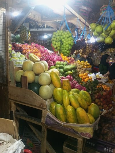 This image depicts a vibrant fruit stall located within a market in Lê Thanh Nghị Ward, Vietnam. The foreground and midground are dominated by a dense arrangement of fresh fruits on wooden display structures.
In the immediate foreground, a wooden crate holds a large pile of ripe yellow-green papayas. To their left, a substantial quantity of spherical, light green and yellowish melons, resembling cantaloupes or honeydew, are stacked, with one melon slice prominently displayed on top. A dark green watermelon is partially visible beneath the melon pile.
Further into the display, various other fruits are arranged: bright pink dragon fruits, clusters of green mangoes, and mounds of small orange citrus fruits, likely oranges or tangerines. Green apples, individually wrapped in white protective netting, are visible on a shelf to the left. Red, spiky fruits, consistent with rambutans or lychees, are also present.
Overhead, numerous fruit bunches hang from the market's wooden ceiling structure by blue ropes. These include large clusters of green grapes, green-yellow pomelos or grapefruits, dark purple fruits (possibly dark grapes), and a textured brown fruit. A single pineapple hangs suspended to the far left. The market stall features exposed wooden beams, metal mesh, and multiple bright artificial lights illuminating the produce.
In the background, several individuals are present. One person, wearing a dark green top and a black head covering (hijab), stands to the right, partially obscured by the fruit displays. Another person with a dark head covering is visible further back. A man in a dark shirt is positioned behind a section of the fruit display. The scene indicates commercial activity in an indoor or covered market setting.
