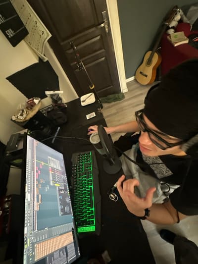 An overhead view depicts a young individual seated at a desk in an indoor environment in Marcoussis, France, actively engaged with a computer setup. The person wears a dark beanie, black-rimmed glasses, a light grey shirt with a dark patterned fabric underneath, and a dark outer garment, along with over-ear headphones. Their right hand operates a black computer mouse, while their left hand is raised and slightly curled, positioned near a black cylindrical microphone equipped with a pop filter.

The desk surface is dark. A large vertical computer monitor on the left displays a Digital Audio Workstation (DAW) interface, showing multiple audio tracks and waveforms. To the right of the monitor is a black gaming keyboard with green backlighting. A speaker or audio equipment is visible to the left of the monitor. Further back on the desk, near the wall, are a white rectangular dish with contents, a small remote control, and packaged items. A fishing rod is leaning against the wall, extending upwards from the desk area.

The background includes a white wall on the left adorned with a black letter board displaying "SAY: SUN" and a white sheet with musical notation. Below these, a black acoustic panel is affixed to the wall. A dark brown wooden door is visible in the center-background, slightly ajar. To the right of the door, a dark teal-green wall is visible, against which an acoustic guitar leans. A dark red armchair or furniture piece next to the guitar holds several books and a light-colored object. The floor is covered with light grey wooden or laminate flooring. A dark green piece of fabric lies on the floor near the door.