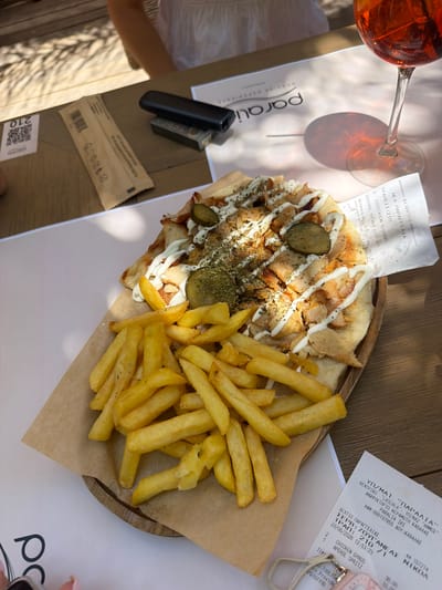 A close-up, overhead shot captures a meal of chicken gyros and fries served on a wooden platter, resting on a light-colored surface. To the right of the food is a receipt from a establishment named "PARALIA," suggesting a location in Paralia, Greece. The receipt details a "CHICKEN GYROS" and "APEROL SPRITZ," dated 28/06/2026. A glass with an orange beverage, likely the Aperol Spritz, is visible in the upper right corner. A person's hand with pink-painted fingernails is partially visible on the left side of the frame. In the background, a person is seated, wearing a white garment. The table appears to be wooden, with some shadows cast from overhead lighting. Additional items on the table include a vape device and a wrapped item with a barcode. The scene is well-lit, suggesting an outdoor or brightly lit indoor setting.