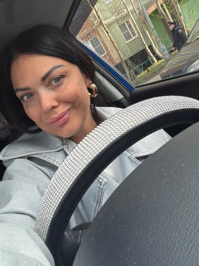 A woman with dark hair and blue eyes is inside a car, her face visible behind a steering wheel covered with rhinestones. She is wearing a light-colored denim jacket and a leopard-print earring. Through the car window, buildings are visible, suggesting an urban environment. Based on the location provided (Murmansk, Russia), the surrounding buildings might reflect architectural styles common in Russian cities, although the details are limited by the viewing angle. A person is vaguely visible outside of the car.