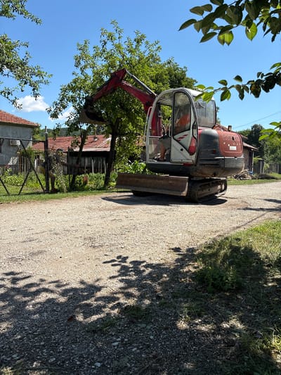 In Oreshak, Bulgaria, on a sunny day, a red and white mini-excavator sits on a gravel path. The excavator's cab is open, revealing a person inside wearing an orange shirt and dark shorts. The excavator's boom is raised, with the bucket positioned above a patch of greenery to the left. In the background, traditional Bulgarian houses with red tile roofs are visible, surrounded by trees and a wire fence. A person, wearing a dark t-shirt and yellow shorts, is visible behind the fence on the left, holding a branch. The scene suggests ongoing work or construction in a rural, residential setting.