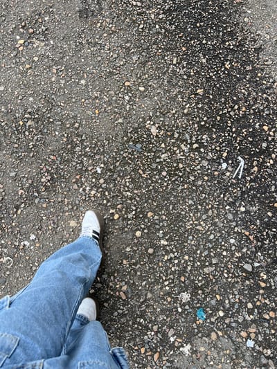 A first-person perspective shot shows a person's legs and feet standing on a wet, pebbled surface, likely asphalt or concrete. The person is wearing light blue jeans and white sneakers with black stripes. The ground is speckled with various sized pebbles and some small pieces of litter, including a white plastic straw and some blue and white plastic fragments. The lighting suggests it is daytime, and the wetness of the ground indicates recent rain or damp conditions. The scene appears to be an outdoor, possibly urban or semi-urban, environment given the litter. The overall impression is casual and ordinary, with no specific activity or strong emotion conveyed beyond the simple act of standing. No visible text is present in the image.