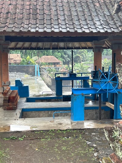 This image depicts an outdoor scene, likely in a rural or semi-rural area of Klungkung, Indonesia, given the tropical vegetation and traditional architecture visible in the background. The foreground is dominated by a large, bright blue mechanical structure, possibly part of a water control system or irrigation equipment. This structure features a large steering wheel, gears, and supporting beams, all painted in a vibrant blue.

To the left of the blue machinery, there is a section of a building with a roof made of dark, curved tiles, characteristic of Indonesian architecture. Beyond this building, a body of water flows, leading towards a small waterfall or dam with a blue railing and steps. In the background, several traditional Indonesian houses with thatched or tiled roofs are nestled amongst lush green trees, including palm trees, suggesting a warm and humid climate. The overall lighting suggests it is daytime, likely overcast or early morning/late afternoon due to the diffused light.

The ground around the machinery is a mix of concrete and muddy earth with some small green plants growing. A stack of wooden planks is visible near the bottom right corner, possibly for construction or repairs. There are no people visible in the image. The scene appears to be calm and functional, showcasing infrastructure within a natural, verdant environment. No discernible text is visible in the image.
