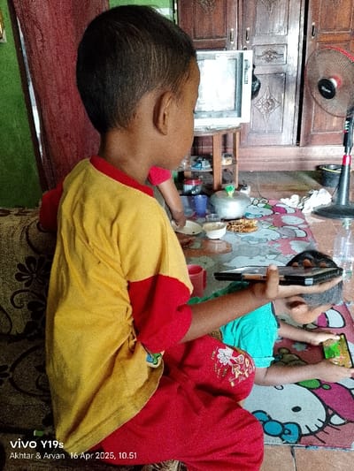 Two young children, one wearing a yellow shirt with red trim and red pants, and one wearing a green shirt, are seated on a floor covered with a colorful patterned rug. The child in the yellow shirt is holding a black tablet and looking at it. The child in the green shirt is lying on the floor with his face turned away from the camera, and looking at a smartphone.  The scene appears to be inside a home in Anjatan, Indonesia. The home features wooden cabinets, a television, and a fan. There is a small bowl of food and a cup on the floor, and a few other items of furniture are visible in the background. The image was taken with a smartphone camera and the date and time are recorded as "16 Apr 2023, 10.51".