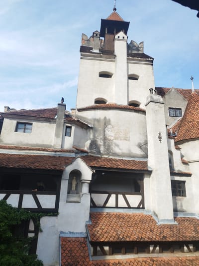The image shows a view of Bran Castle, also known as Dracula's Castle, located in the city of Predeluț, Romania. The castle is a medieval fortress with a white, multi-story structure characterized by red tile roofs, tall chimneys, and numerous windows. In the foreground, several layers of red tiled roofs are visible. The middle ground features a white wall with a statue in a niche, an arched entryway covered in ivy, and a balcony with wooden crossbeams. The background is dominated by the imposing white main tower of the castle, topped with a dark, stepped roof and a small spire. The sky is partly cloudy and blue. A person is visible on the balcony.