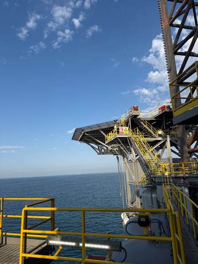 An elevated perspective from a platform, possibly part of an offshore oil rig, showcases a vast expanse of the Mediterranean Sea under a bright blue sky dotted with white clouds. The foreground is dominated by a bright yellow safety railing. Behind the railing, a complex metal structure with multiple levels, staircases, and walkways is visible. This structure is painted in a utilitarian grey and black, with yellow accents on railings and stairs for visibility. Various industrial components and equipment are present on the platforms. The sea stretches to the horizon, where faint outlines of distant landmasses or structures can be seen. The scene appears to be a clear, sunny day.