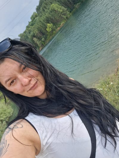 In the city of Dobrotino, Bulgaria, a woman with long, dark hair takes a selfie outdoors. She is wearing a white tank top and sunglasses on her head. Her left arm, with a tattoo, is visible in the foreground. Behind her, a body of water, likely a lake or reservoir, is visible, with trees lining the opposite shore. The sky is overcast.
