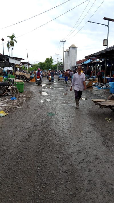 The image depicts a bustling outdoor market scene in Geulanggang Teungoh, Indonesia, under an overcast sky. The ground is predominantly unpaved and muddy, with numerous puddles and debris scattered across it, suggesting recent rainfall. Several people are present, engaged in various activities. In the foreground, a man wearing a light-colored shirt and dark pants, along with tall grey rubber boots, walks towards the camera. Behind him, a woman on a motorbike with a pink headscarf is visible. Further down the lane, more individuals can be seen navigating the market. The market stalls are constructed with dark roofing materials, and some are illuminated by overhead lights. Objects like carts, motorcycles, and various containers, possibly for goods, are dispersed throughout the scene. In the background, palm trees and what appear to be tall, white structures with domed tops, resembling minarets, are visible. The overall atmosphere suggests a busy commercial environment, likely a fish market given the presence of what looks like fish laid out on tables in the background.