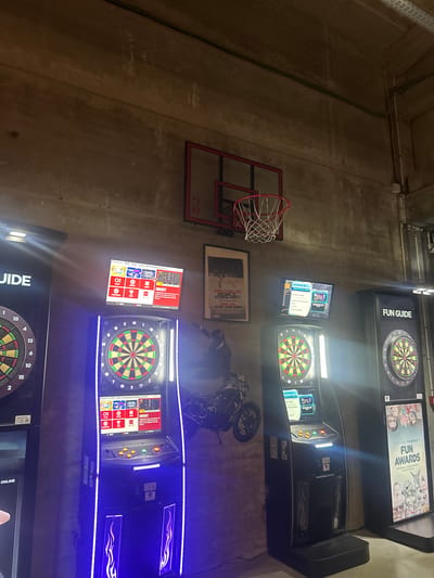 This media file captures an indoor recreational space, likely an arcade or bar, with a strong emphasis on electronic dart games. The scene is dominated by multiple, brightly lit dart machines with prominent blue LED accents, set against a backdrop of concrete walls.
In the foreground, a partial view of an electronic dart machine shows a dartboard with numbers 20, 1, 18, 4, 13, 6, 10, 15, and 2. Above it, a screen displays "JIDE" and "ONLINE." Further back, two more dart machines are visible. The top one features a screen with "FUN GUIDE" next to a dartboard and another screen above it displaying "FUN AWARDS" with images of people and the text "Animations en temps." The middle machine has a screen showing game options like "CRICKET" and "501," along with "OPTION DE JEU" and a website address "phoenixdarts.com.fr."
To the left of the dart machines, a miniature red basketball hoop with a net is affixed to the concrete wall. Below the hoop and positioned between it and the dart machines, a framed poster advertises a "PYSAMA PARTY" (Pajama Party) scheduled for "SAMEDI 21 MARS 2026." Other French text on the poster includes "BEGANES" and "SET CONSO OFFERTE." A dark motorcycle with visible wheels and handlebars is parked between the middle and bottom dart machines. The overall lighting is dim, relying heavily on the illumination from the electronic machines, creating an evening or nighttime atmosphere. The concrete walls and exposed wiring at the top suggest a functional, possibly industrial, setting for entertainment. The French text throughout confirms the location context of Villeneuve-d'Ascq, France.