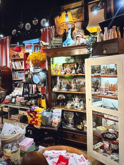 This media file captures a cluttered shop interior, possibly an antique or curio shop, filled with a diverse array of objects. The scene is densely packed with items, suggesting a place where many trinkets and collectibles are displayed.
Prominently featured are shelves and cabinets brimming with porcelain items, including teacups, saucers, and decorative vases, many adorned with intricate patterns in red and white. There are also vintage lamps with shades, and what appear to be framed artworks or prints. In one section, a collection of small, model buildings is visible, set against a backdrop that resembles a city scene. Elsewhere, decorative plates are hung in a line. A spotlight fixture is positioned above a collection of books and other items. In the foreground, a woven basket contains what looks like postcards or letters, and a large tin can is also visible. The overall impression is one of abundance and perhaps a touch of nostalgia.
Visible text on some items includes: "LOVE", "CHEF", and "Новинки". The lighting suggests an indoor setting, possibly during the day or with artificial lighting creating a warm ambiance. No people are visible in the frame, and there are no indications of weather. The presence of text in Cyrillic script, specifically "Новинки", suggests a location in Russia, consistent with the provided location context. The sheer volume and variety of items imply a rich collection of curiosities.