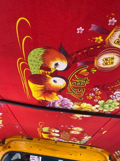 The image displays the patterned interior roof lining of a vehicle, located in Katcha, Nigeria. The lining is a red fabric adorned with a repeating ornate design. This design prominently features two stylized, colorful birds, resembling mandarin ducks, with plumage in green, orange, yellow, white, and black, positioned facing each other. Interspersed within the pattern are various floral elements, including peonies and smaller blossoms rendered in purple, pink, yellow, and green hues. Chinese characters are also integrated into the design, specifically the "Double Happiness" symbol (囍) appearing on decorative lanterns, and a character resembling "Spring" (春). A black rubber trim runs horizontally across the lower portion of the image, separating the patterned fabric ceiling from the vehicle's yellow-painted metal frame. An unidentifiable element, possibly a reflection or a small poster, is partially visible within the yellow frame on the lower left.