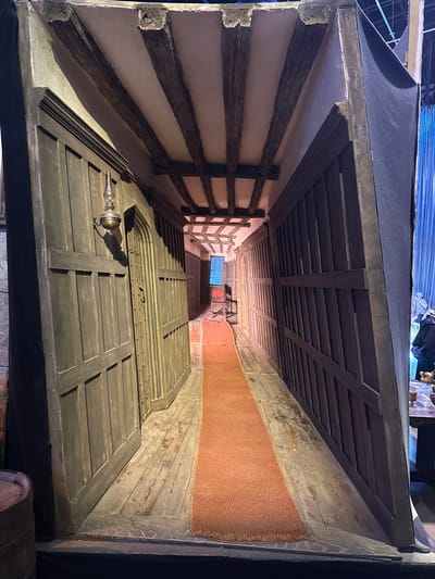 This media file shows an interior scene, likely a hallway or corridor. The perspective is from a high angle, looking down into the space. The architecture suggests an older building, with exposed wooden beams and a planked ceiling. The walls are textured and appear to be plastered or stuccoed. A reddish-brown carpet or runner runs along one side of the corridor.
Dominating the foreground is a large, dark wooden door with a grid-like pattern created by intersecting planks. A metal fixture, possibly a door knocker or handle, is visible on this door. Further down the corridor, a small, multi-paned window is visible, letting in a soft blue light, suggesting daylight, though the overall lighting in the scene is dim and atmospheric. The scene is constructed to evoke a historical or perhaps a fantasy setting, possibly a film or theatre set. There are no people visible in the frame. The overall impression is one of depth and age, with a somewhat mysterious or imposing atmosphere. The location context of Watford, United Kingdom, does not directly inform the visual elements of the scene itself, but it is provided as external information.