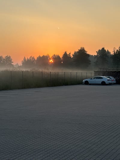 A sunrise scene is captured, with a warm orange glow filling the sky. The sun is partially obscured by trees and a layer of fog, creating a hazy effect. In the foreground, a paved parking lot is visible, with white lines marking parking spaces. Two white cars are parked in the lot, facing away from the camera. A fence runs along the edge of the parking lot, separating it from a grassy area and trees. The overall atmosphere is serene and tranquil.